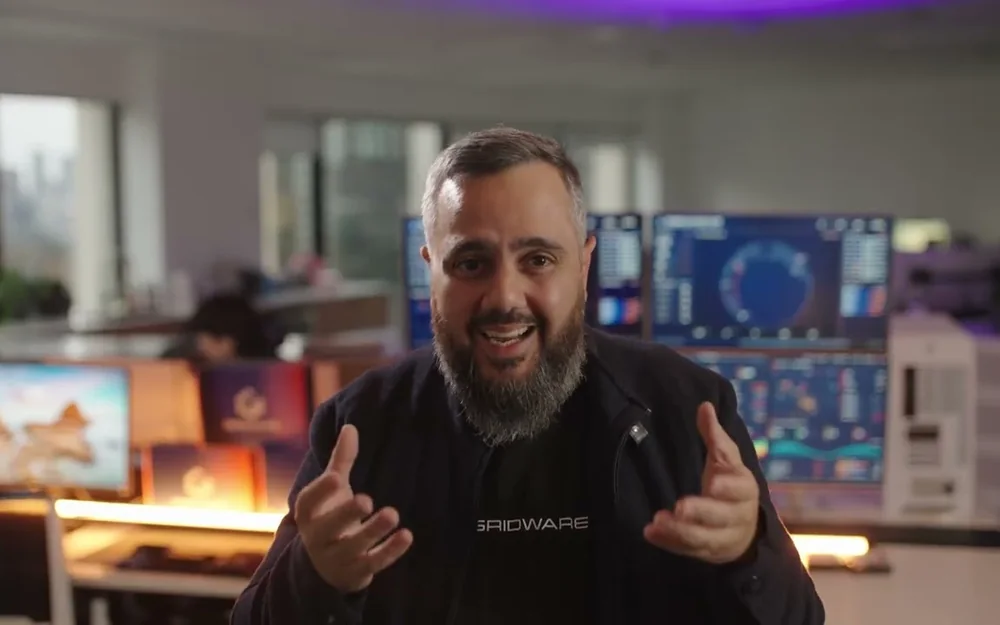 Ahmed Khanji speaking to camera in the Gridware office, with security monitoring screens behind him.