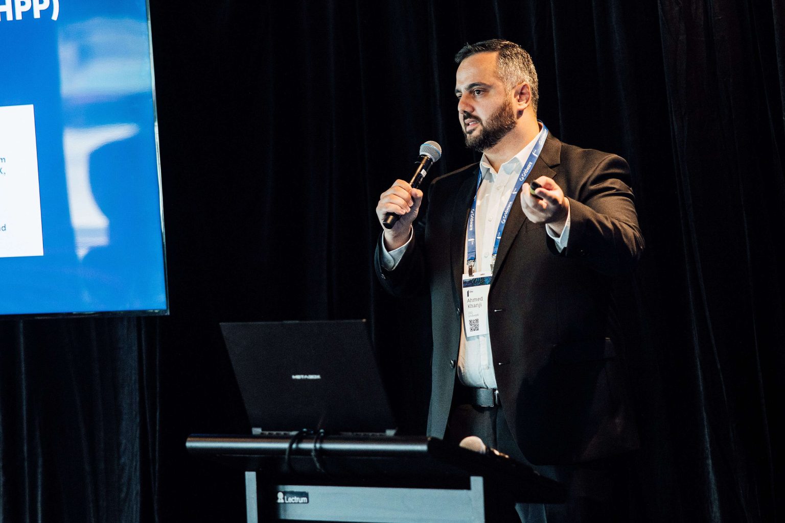 Ahmed Khanji, CEO of Gridware, presenting on stage with a microphone and a Gridware conference lanyard.